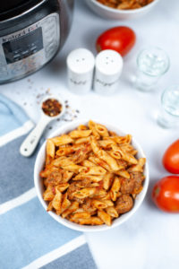 White bowl of penne vodka sauce with tomatoes and seasonings on the side.