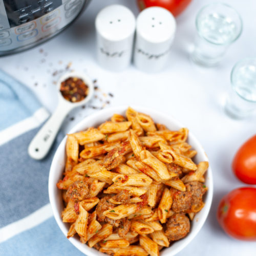 White bowl of penne vodka sauce with tomatoes and seasonings on the side.