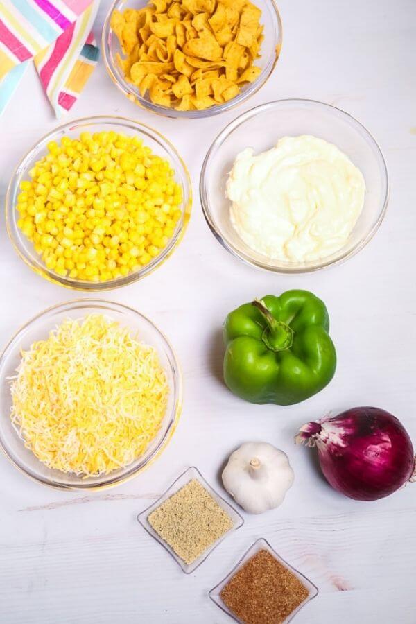 Various ingredients arranged on a table for Frito Salad, including corn, shredded cheese, bell pepper, red onion, garlic, seasonings, and chips.