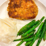Round white plate with breaded pork chops, green beans and mashed potatoes.