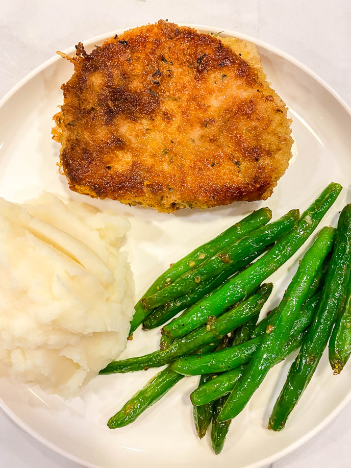 Round white plate with breaded pork chops, green beans and mashed potatoes.