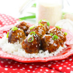 Plate of meatballs and rice on a red and white checked tablecloth with a glass of milk net to it.