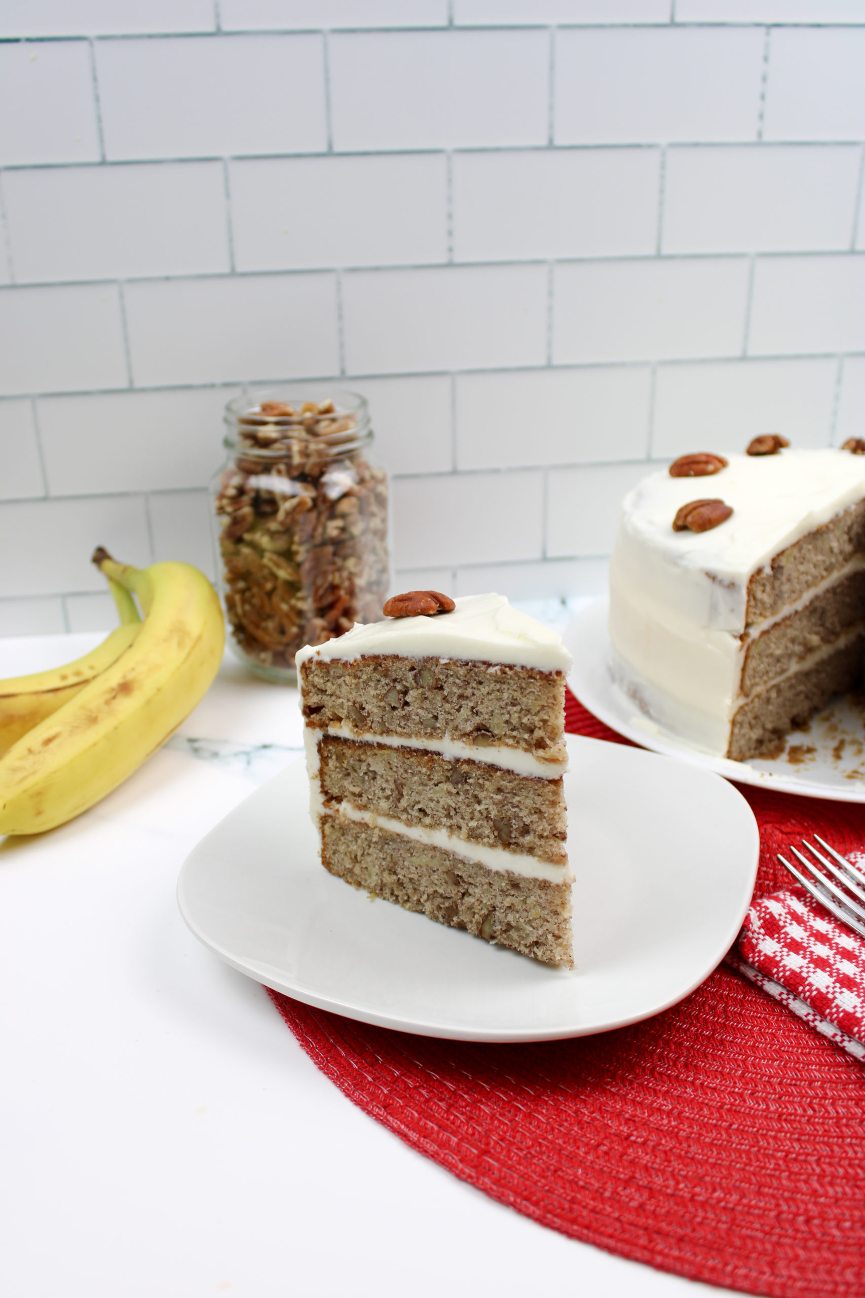 Old Fashioned Hummingbird Cake with a jar of walnuts and a banana in the background.