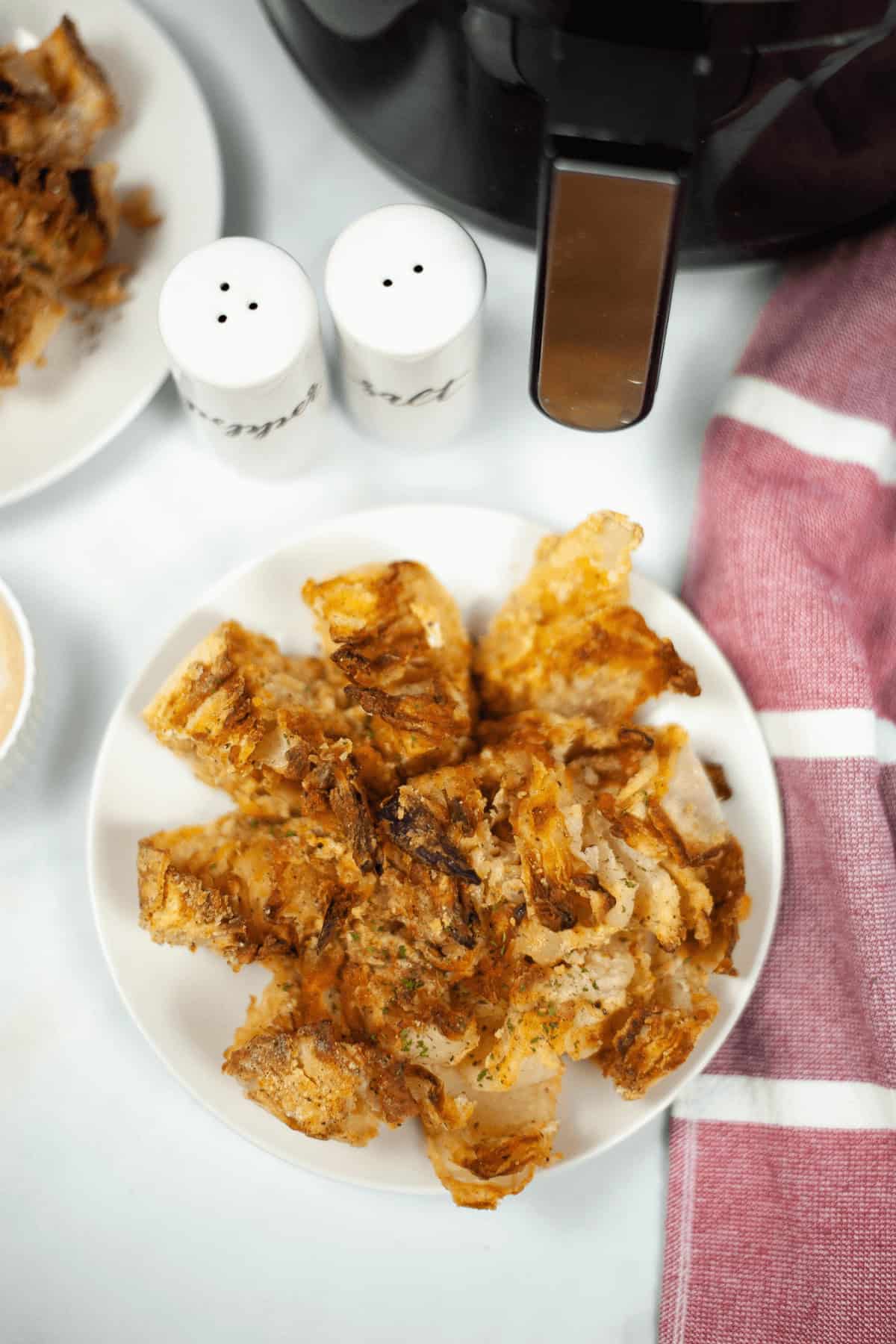 A plate of crispy, golden-brown blooming onion petals sits temptingly next to an air fryer. Salt and pepper shakers accent the scene on a pristine white surface.