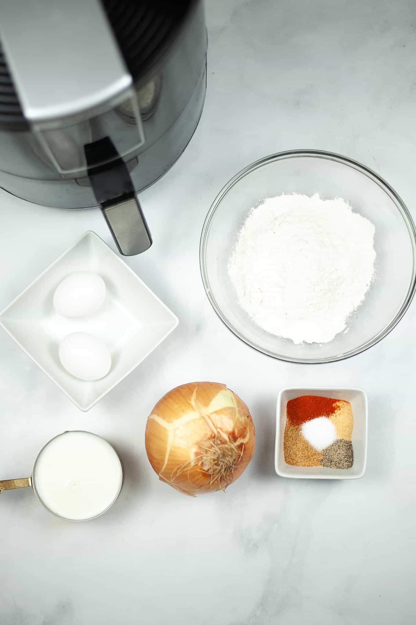 Ingredients for cooking: two eggs, flour in a bowl, and all you need for an Air Fryer Blooming Onion—an onion ready to transform and seasonings in a dish. On the marble surface sits a measuring cup with liquid beside an air fryer, anticipating the creation of deliciousness.