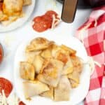A plate of delicious Air Fryer Pizza Rolls sits next to the appliance, surrounded by scattered pepperoni slices and shredded cheese on a sleek white table.