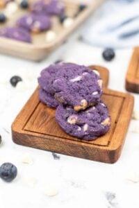 Three blueberry white chocolate cookies with a hint of purple are stacked on a wooden board.