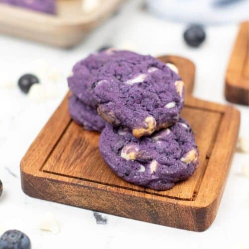 Three blueberry white chocolate cookies with a hint of purple are stacked on a wooden board.
