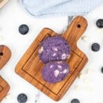 Two blueberry white chocolate cookies studded with white chocolate chips sit on a wooden board, surrounded by fresh blueberries and extra chips. A striped cloth adds a touch of charm to the background.