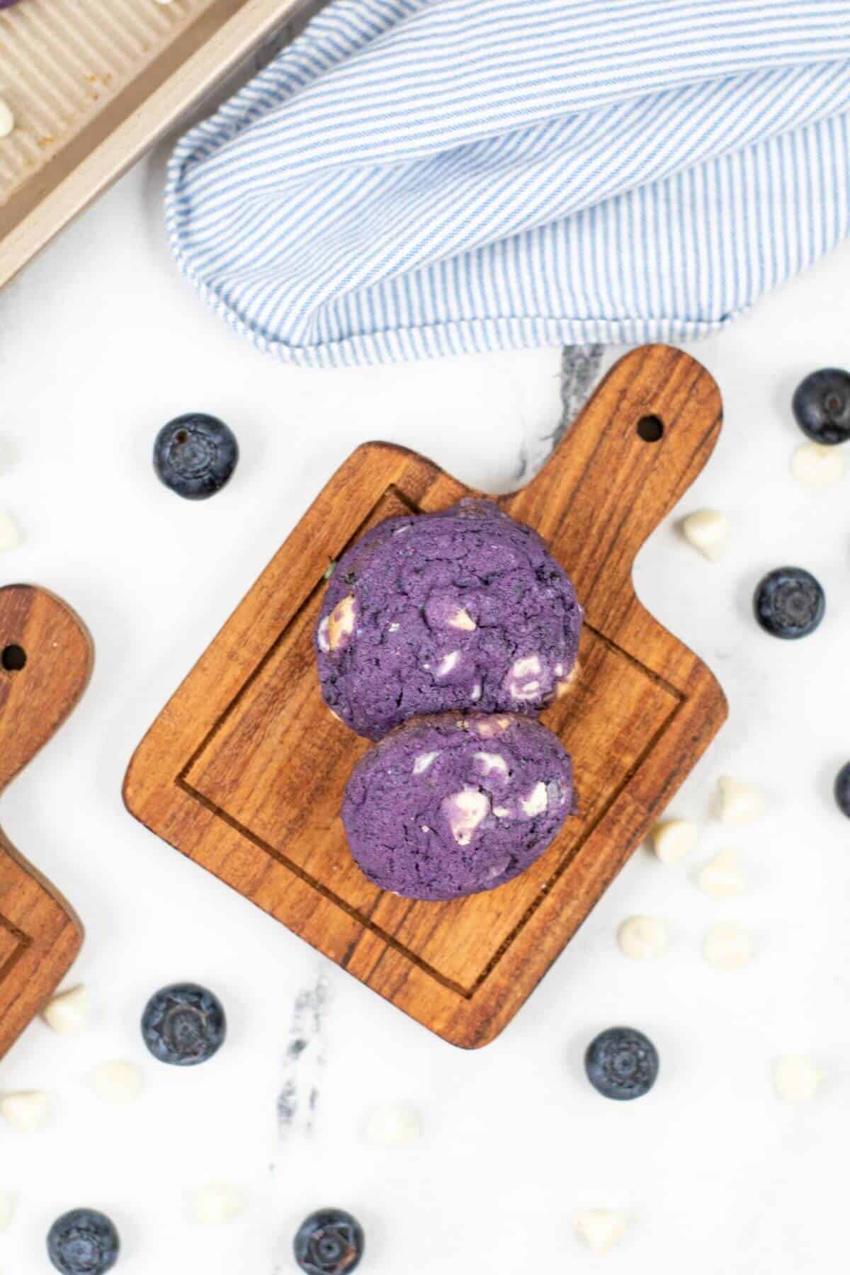 Two blueberry white chocolate cookies studded with white chocolate chips sit on a wooden board, surrounded by fresh blueberries and extra chips. A striped cloth adds a touch of charm to the background.