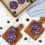 the treats with purple hues rest on wooden boards and a baking tray. They are scattered across the marble surface, while a striped cloth is tucked into the corner.