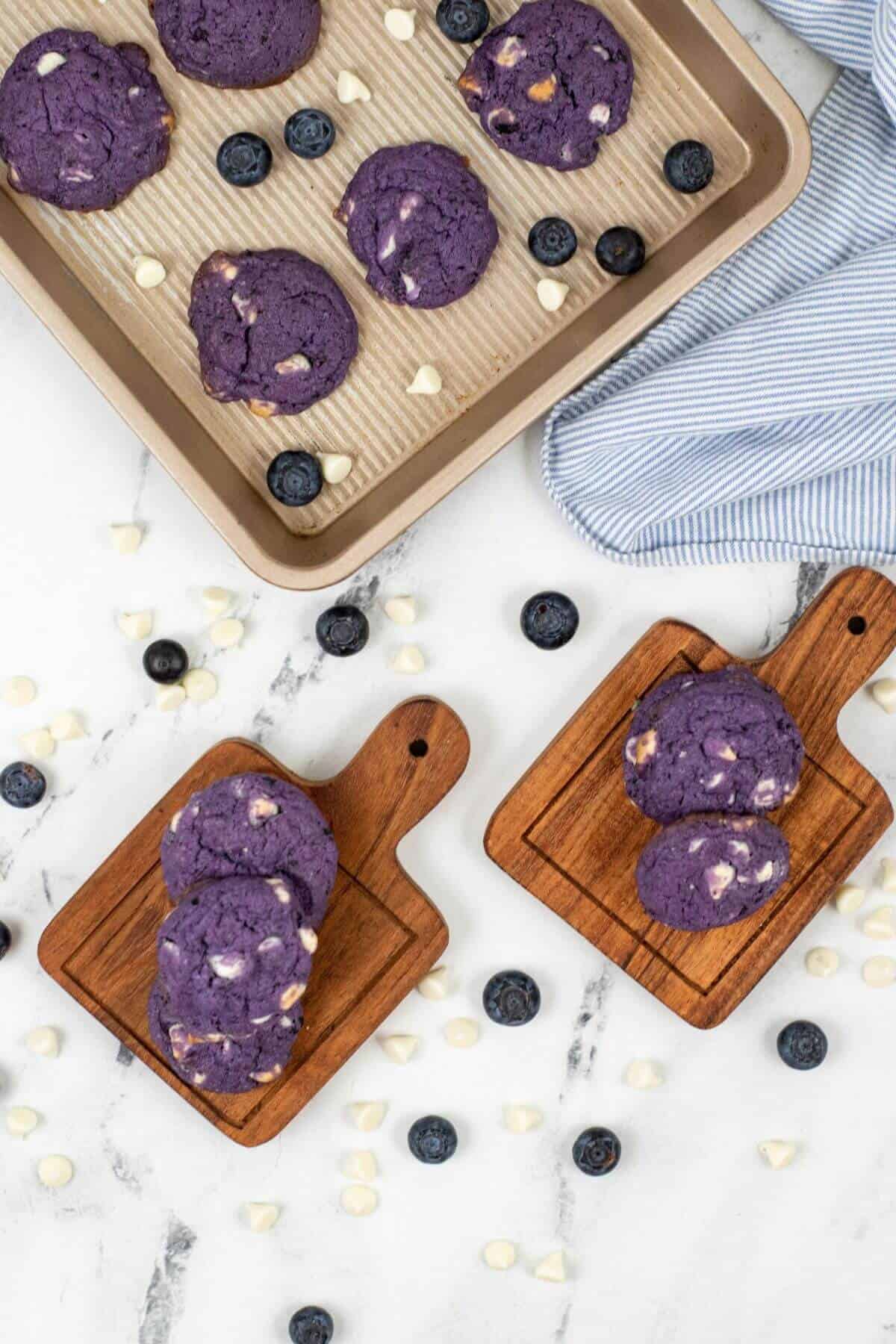 the treats with purple hues rest on wooden boards and a baking tray. They are scattered across the marble surface, while a striped cloth is tucked into the corner.