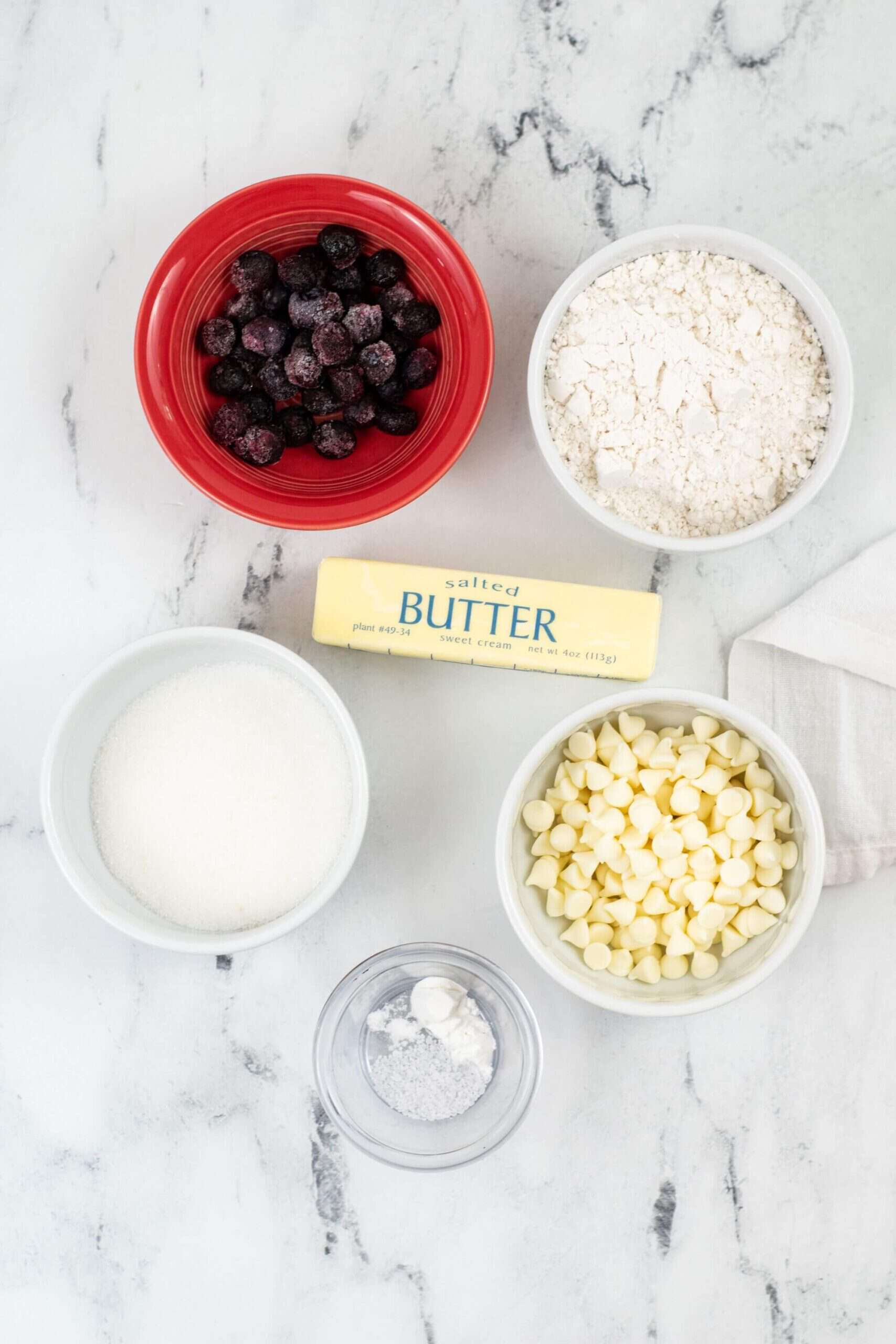 Ingredients for Blueberry White Chocolate Cookies: a red bowl of blueberries, flour in a white bowl, a stick of salted butter, a bowl of sugar, white chocolate chips in a bowl, and a small bowl of baking soda—all on a marble surface.