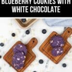 Two wooden boards present blueberry white chocolate cookies, adorned with chunks of white chocolate, surrounded by scattered blueberries and chips on a pristine white surface.