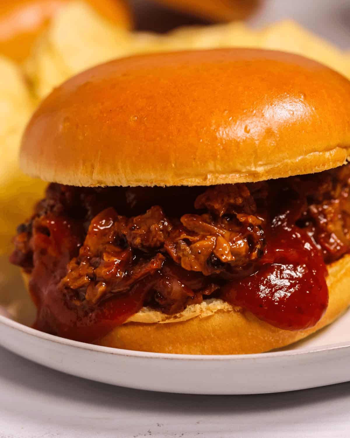 A close-up of a sloppy joe sandwich filled with a rich meat mixture and sauce on a white plate, reminiscent of the flavors found in a chopped brisket sandwich, accompanied by potato chips in the background.