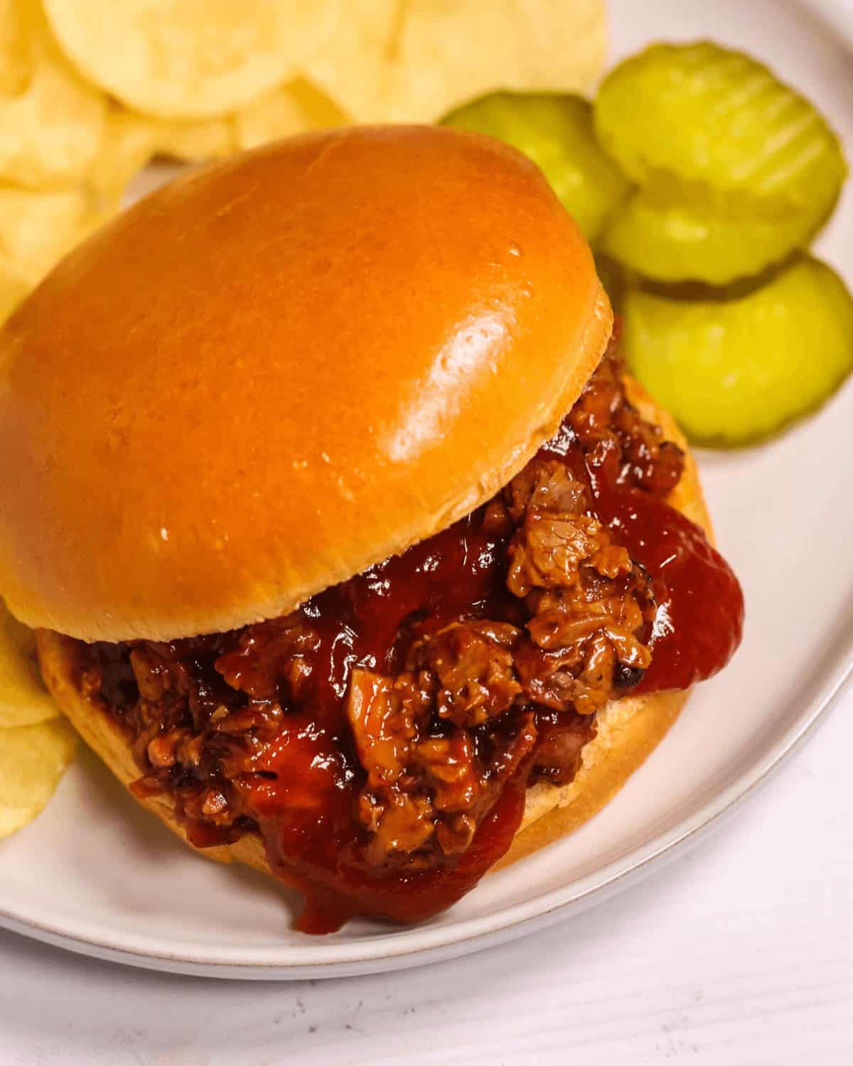 A sandwich with pickles and potato chips on a white plate, offering the comforting taste of a classic chopped brisket sandwich.