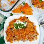 A serving of Dorito Chicken Casserole topped with crushed tortilla chips and cilantro graces a white plate beside a fork, while a dish of the casserole and whole chips linger enticingly in the background.