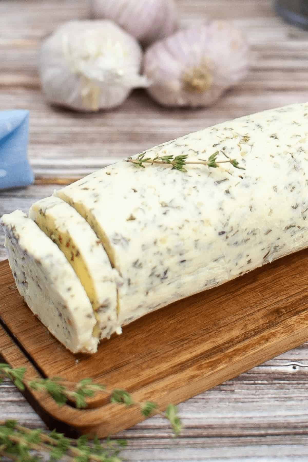 A garlic herb butter log with two slices cut, rests on a wooden board, garnished with fresh herbs. Garlic bulbs linger in the background.