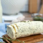 A roll of garlic herb butter rests on a wooden board, with a slice cut from one end and garnished with fresh herbs.
