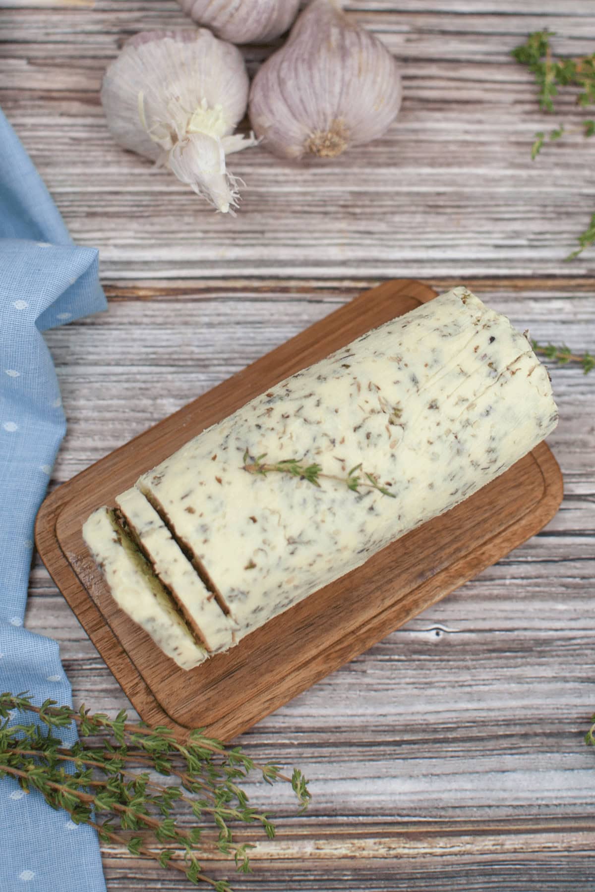 A block of garlic herb butter rests on a wooden board, surrounded by fresh thyme sprigs and garlic bulbs, against a rustic wooden background.