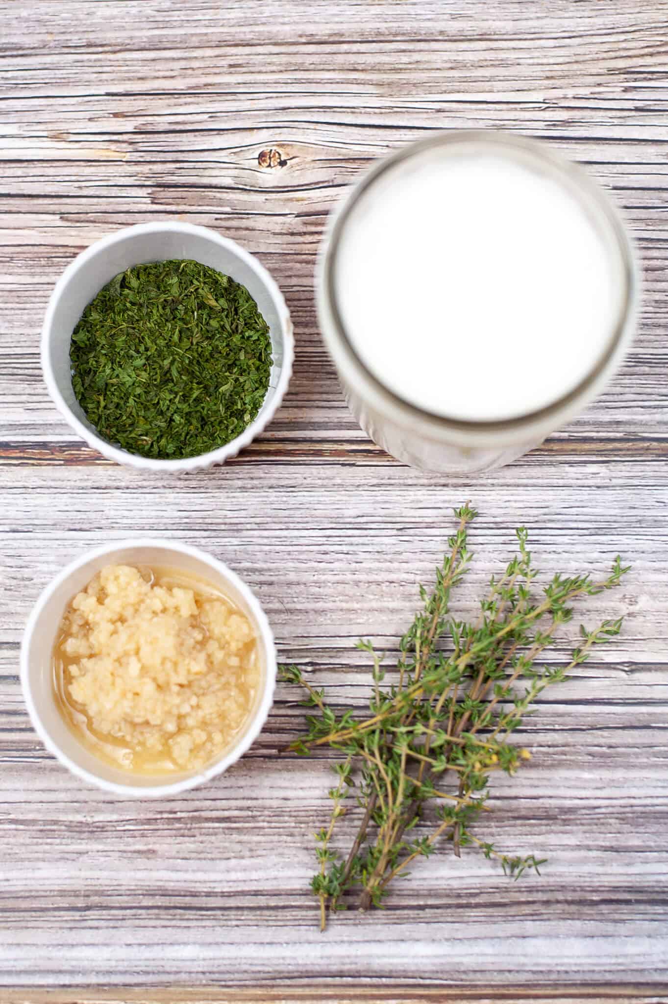 A glass of milk, a bowl of minced garlic, chopped herbs ready for the perfect garlic herb butter blend, and sprigs of thyme grace the wooden surface.