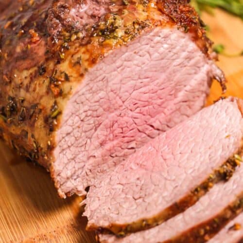 Sliced beef eye of round roast with a seasoned crust, displayed on a wooden cutting board with herbs in the background.