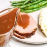 A plate with slices of tender beef eye of round roast topped with brown gravy, mashed potatoes, green beans, and a gravy boat.