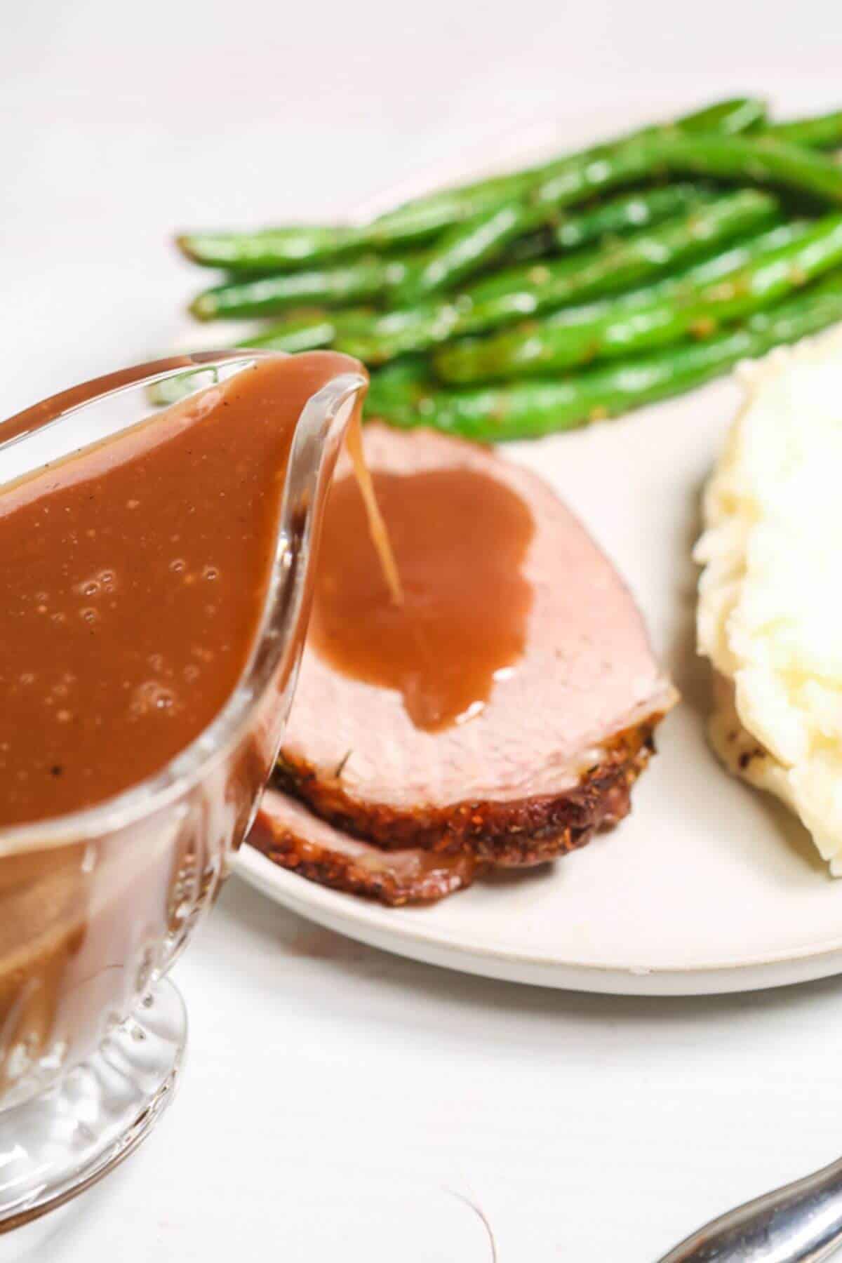 A plate with slices of tender beef eye of round roast topped with brown gravy, mashed potatoes, green beans, and a gravy boat.