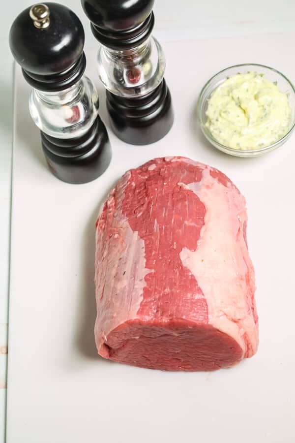 Raw beef eye of round roast on a white cutting board with salt and pepper grinders and a small bowl of herb butter in the background.