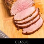 A sliced beef eye of round roast with garlic and herbs is displayed on a wooden cutting board, accompanied by a knife and fresh herbs.