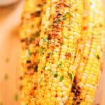 Three grilled Asian corn cobs are stacked on a wooden surface, topped with herbs and a drizzle of sauce. A small dish with dark sauce is partially visible in the background.