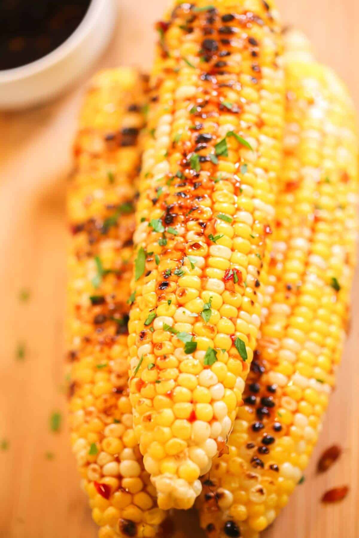 Three grilled Asian corn cobs are stacked on a wooden surface, topped with herbs and a drizzle of sauce. A small dish with dark sauce is partially visible in the background.