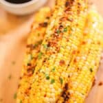 Three grilled Asian Corn cobs topped with herbs and seasoning are stacked on a wooden surface, with a small bowl of dark sauce in the background.