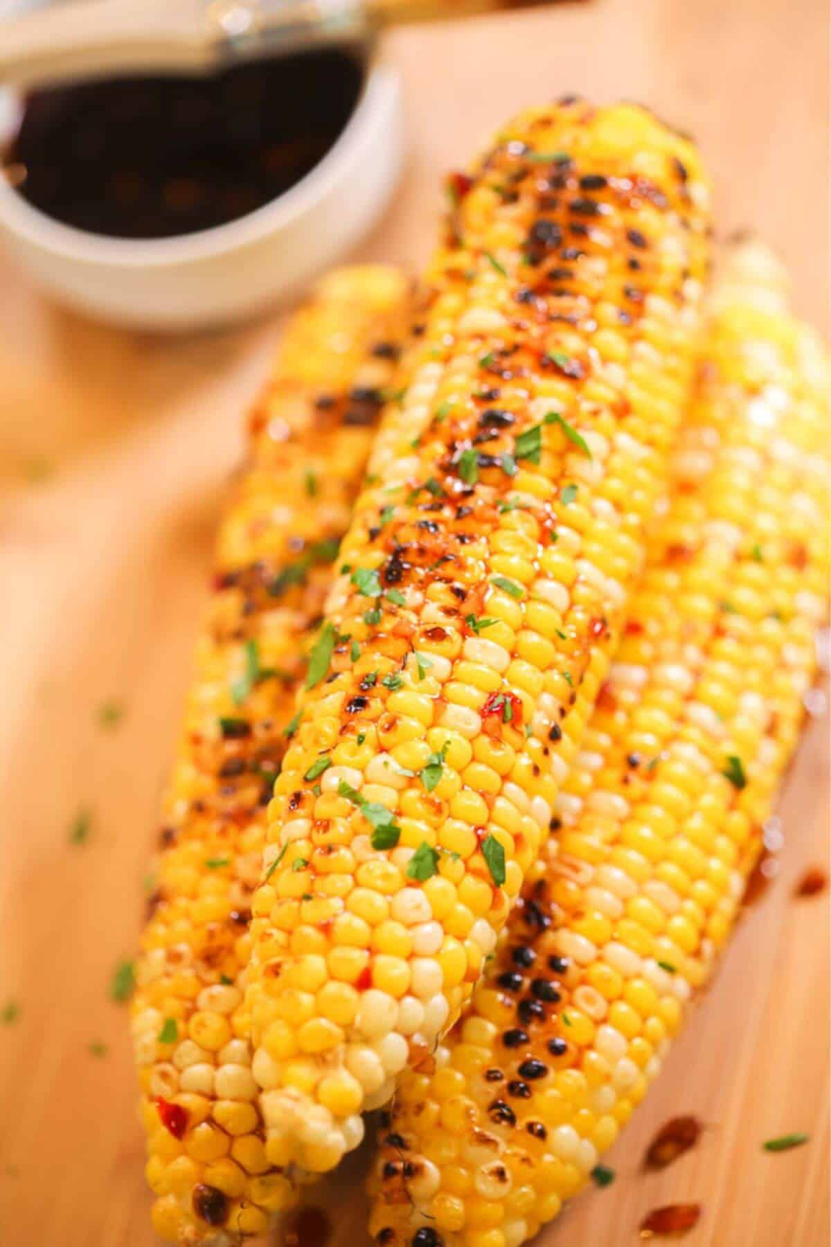 Three grilled Asian Corn cobs topped with herbs and seasoning are stacked on a wooden surface, with a small bowl of dark sauce in the background.
