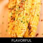 Three ears of grilled Asian Corn rest on a wooden surface, topped with fresh herbs and savory seasoning. Text at the bottom reads: "Flavorful Grilled Asian Corn.