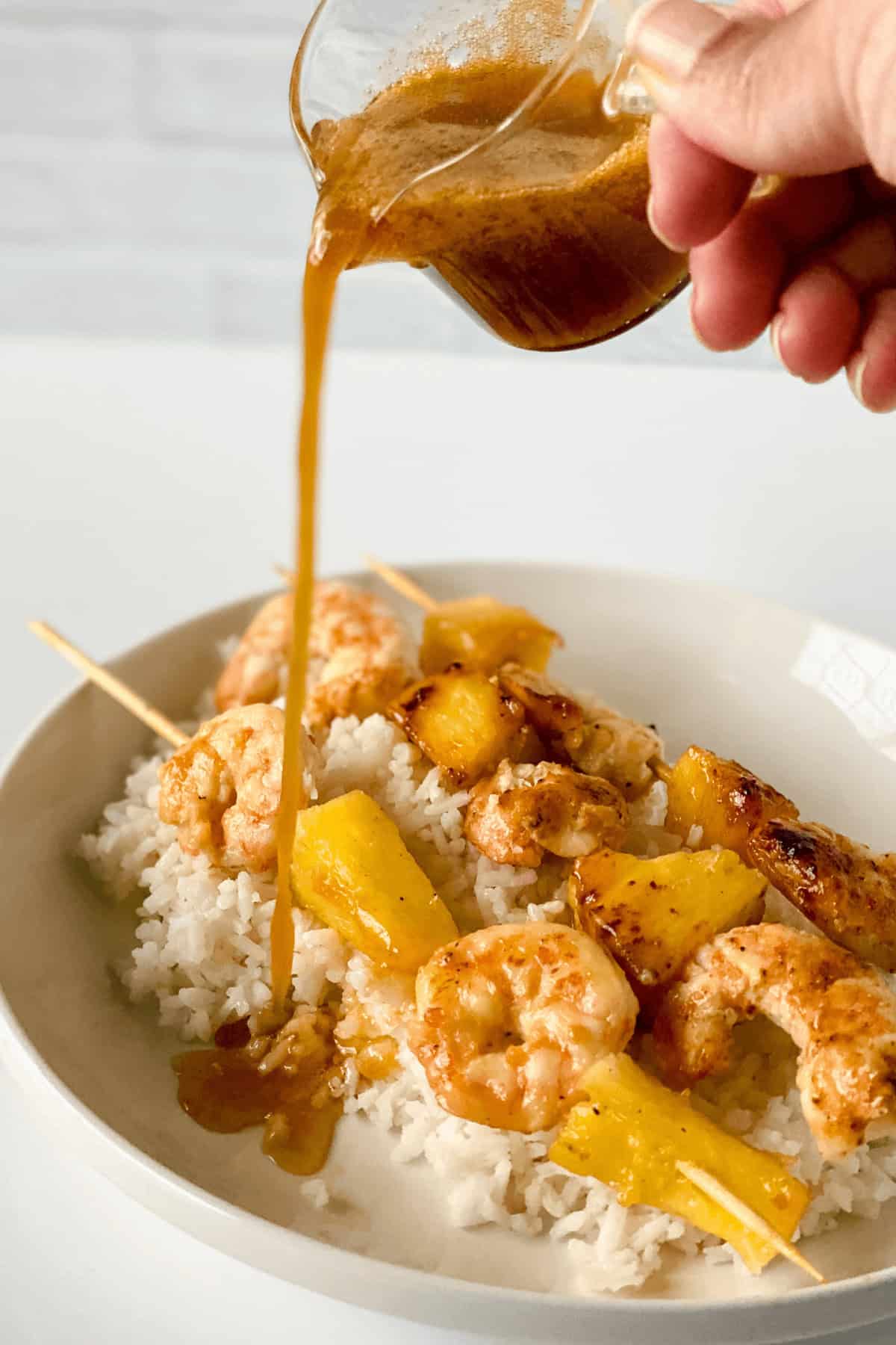 A hand pours sauce over a dish containing Hawaiian Garlic Shrimp Skewers and pineapple, served on a bed of white rice.