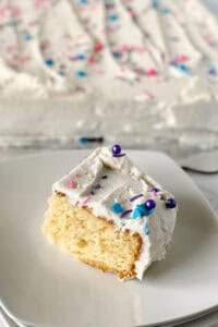 A slice of vanilla sheet cake with white frosting and colorful sprinkles graces a plate, while the rest of the cake sits invitingly in the background.