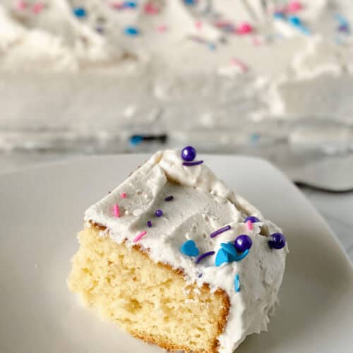 A slice of vanilla sheet cake with white frosting and colorful sprinkles graces a plate, while the rest of the cake sits invitingly in the background.