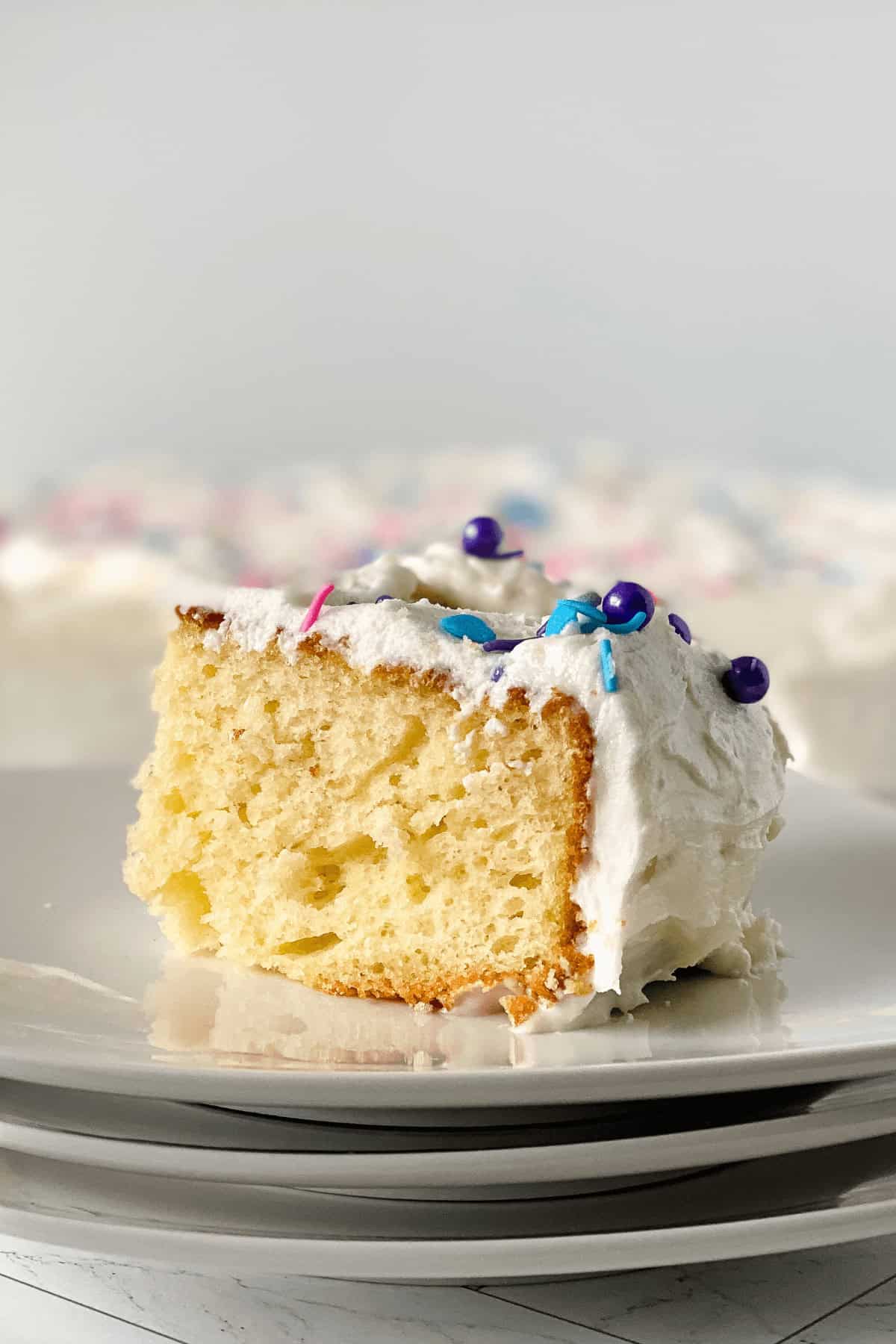 A slice of delicious vanilla sheet cake with white frosting and colorful sprinkles sits atop a stack of plates.