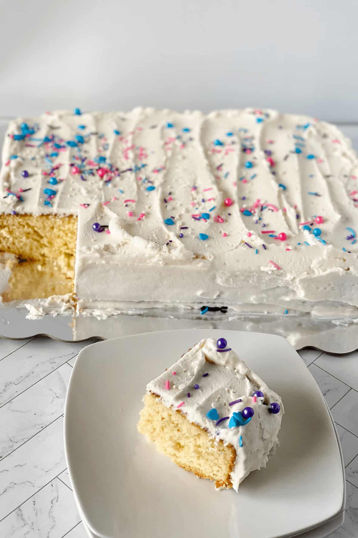 A slice of vanilla sheet cake with white frosting and colorful sprinkles sits on a plate. The remaining cake rests behind it on a serving tray, just waiting to be devoured.