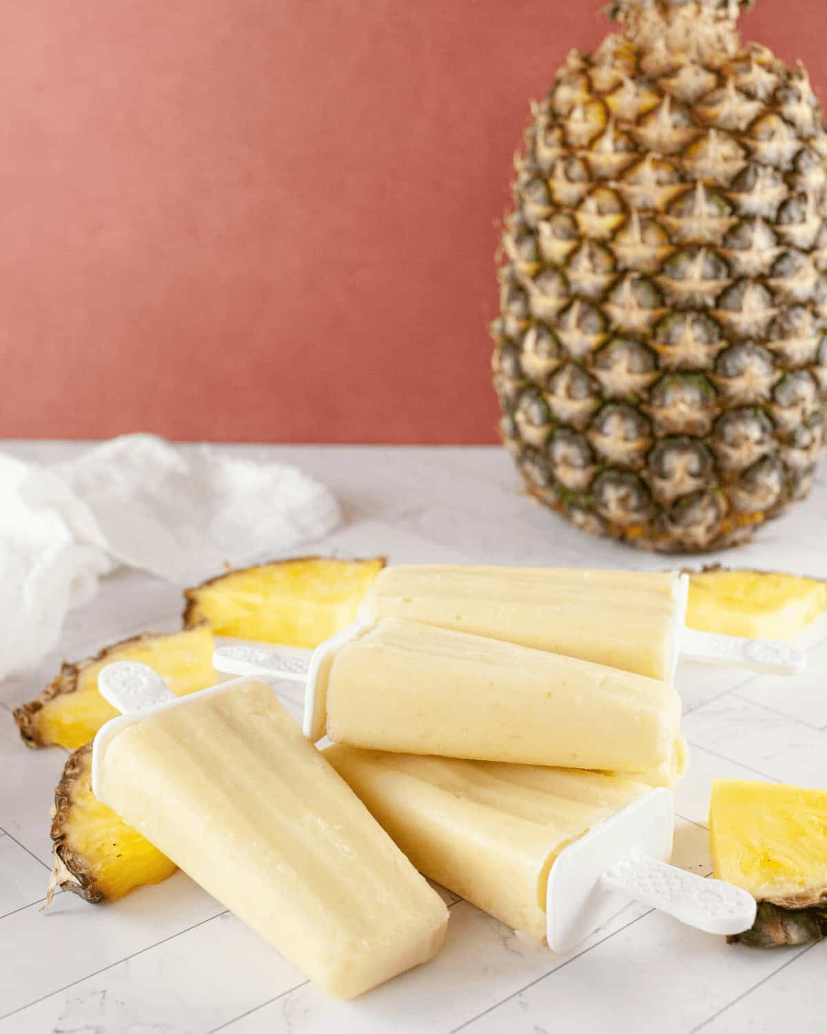 Pineapple Popsicles on a marble surface with fresh pineapple slices and a whole pineapple in the background.