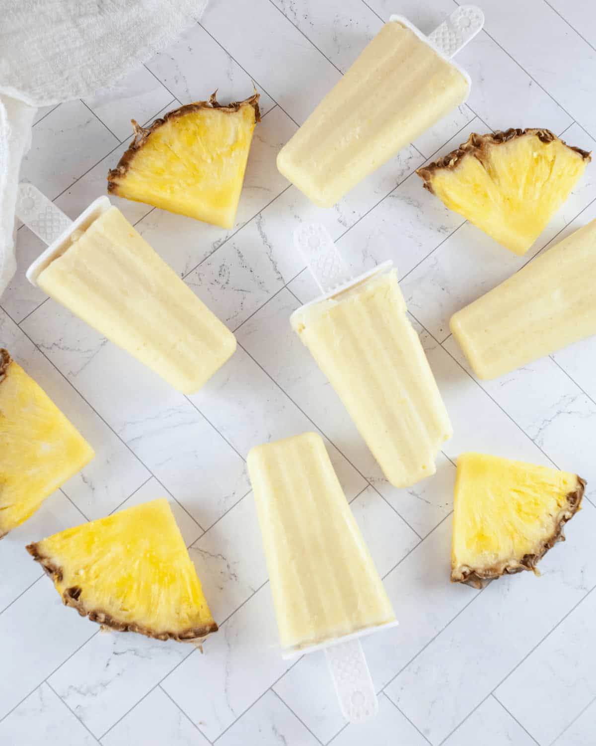 Homemade pineapple popsicles artfully arranged with fresh pineapple slices on a white marbled surface.