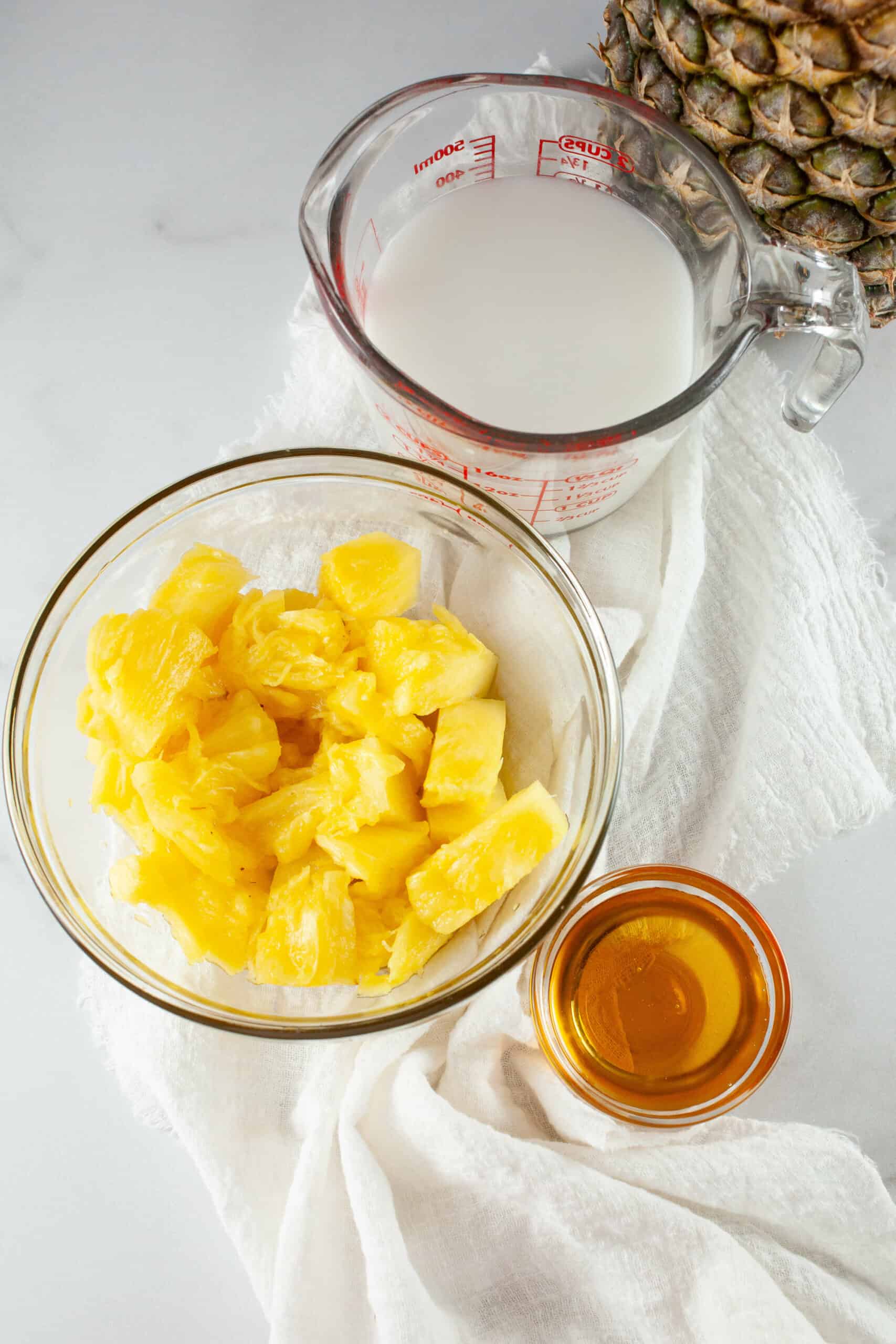 Ingredients for Pineapple Popsicles on a kitchen countertop, featuring chopped pineapple in a bowl, a measuring cup with milk, and a small bowl of honey.