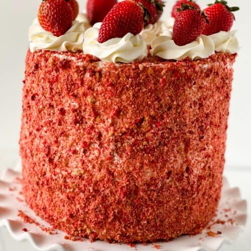 A tall round Strawberry Crunch Cake, made with our delicious Strawberry Crunch Cake Recipe, covered in red strawberry crumbs, topped with swirls of white frosting and whole strawberries, displayed on a white cake stand.
