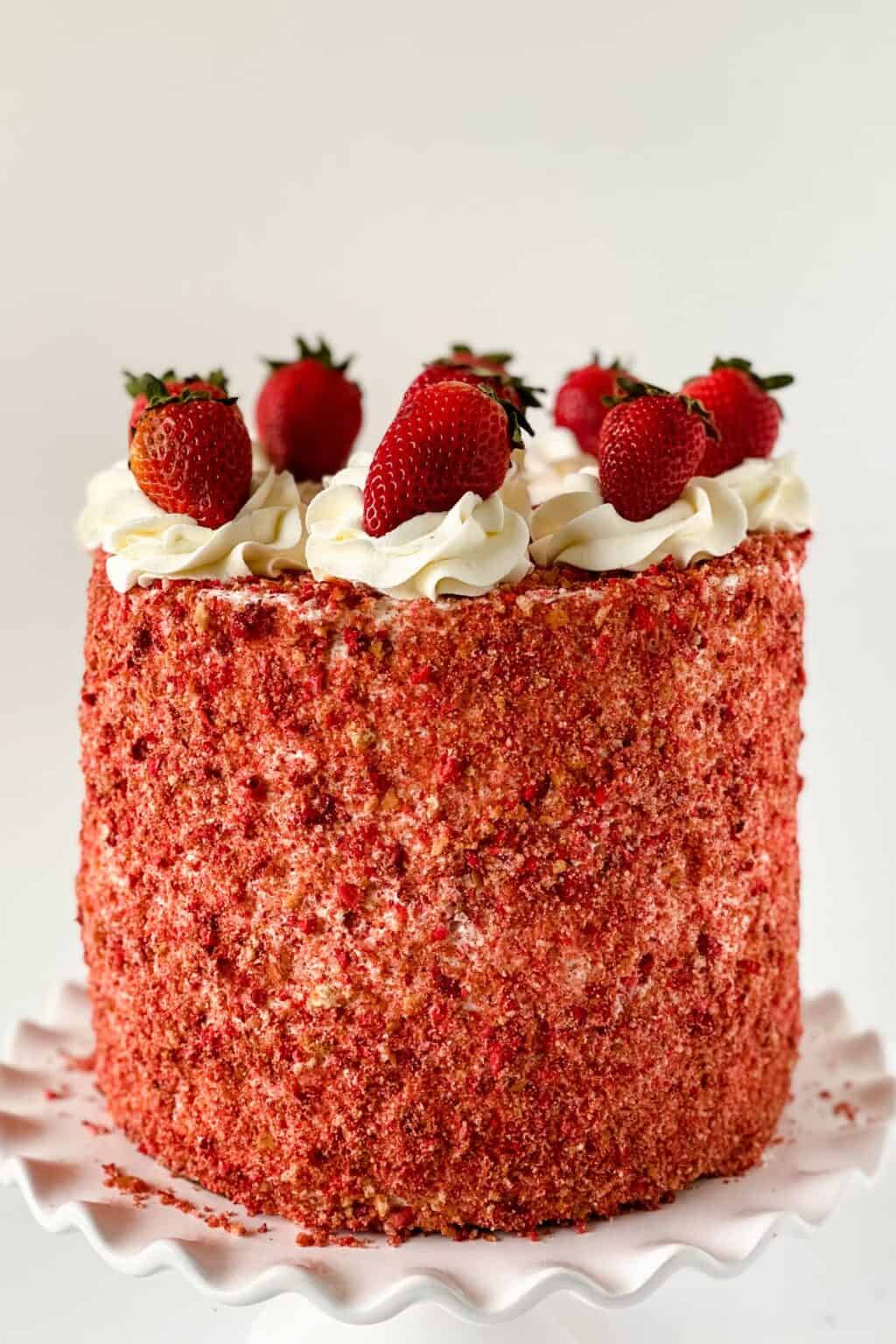 A tall round Strawberry Crunch Cake, made from our delicious Strawberry Crunch Cake Recipe, is covered in red strawberry crumbs, topped with swirls of white frosting and whole strawberries, and displayed on a white scalloped cake stand.