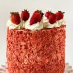 A tall round Strawberry Crunch Cake, made from our delicious Strawberry Crunch Cake Recipe, is covered in red strawberry crumbs, topped with swirls of white frosting and whole strawberries, and displayed on a white scalloped cake stand.