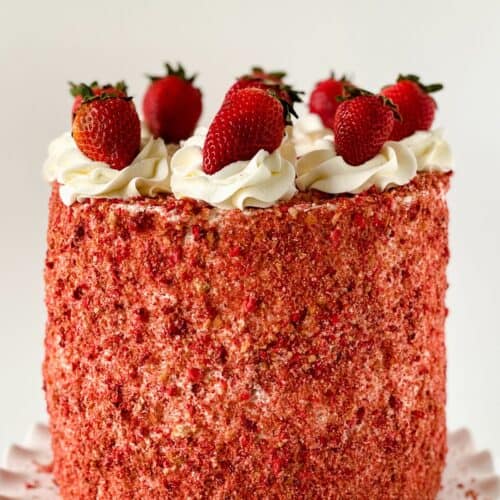 A tall round Strawberry Crunch Cake, made from our delicious Strawberry Crunch Cake Recipe, is covered in red strawberry crumbs, topped with swirls of white frosting and whole strawberries, and displayed on a white scalloped cake stand.