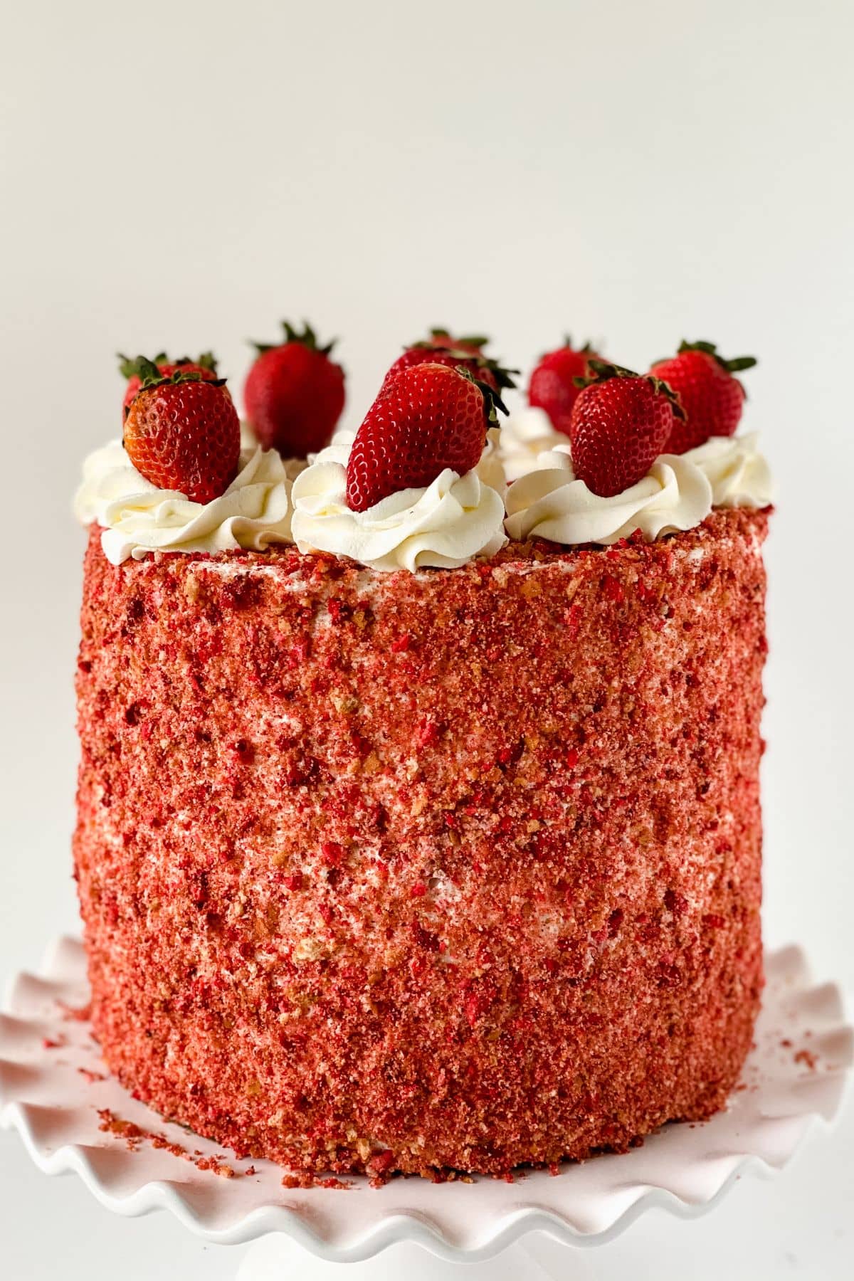 A tall round Strawberry Crunch Cake, made from our delicious Strawberry Crunch Cake Recipe, is covered in red strawberry crumbs, topped with swirls of white frosting and whole strawberries, and displayed on a white scalloped cake stand.