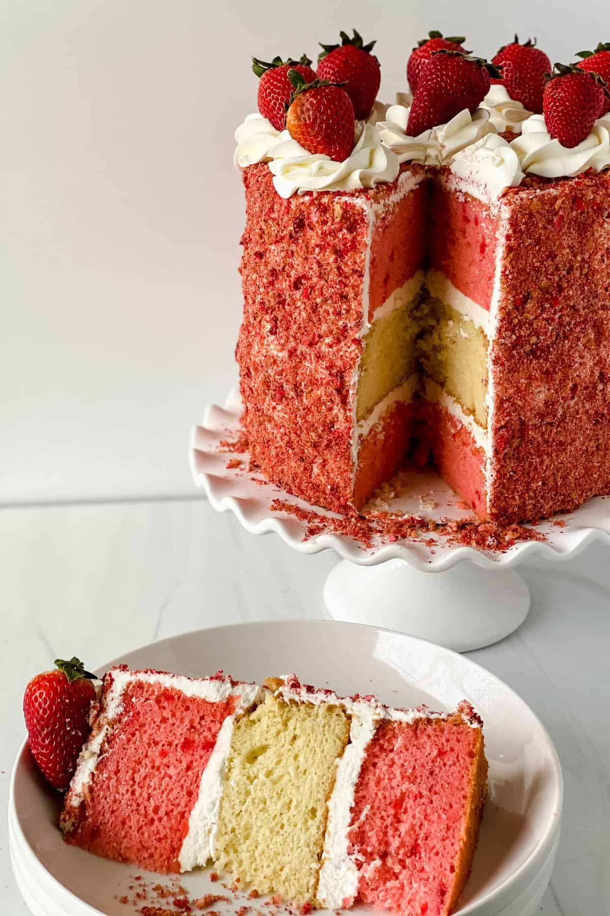 A tall layered cake with pink and yellow sponge, white frosting, and strawberries on top—just like a classic Cake Recipe.
