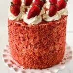 A tall Strawberry Crunch Cake Recipe creation covered in red crumbs, topped with swirls of white frosting and whole strawberries, displayed on a white scalloped cake stand.